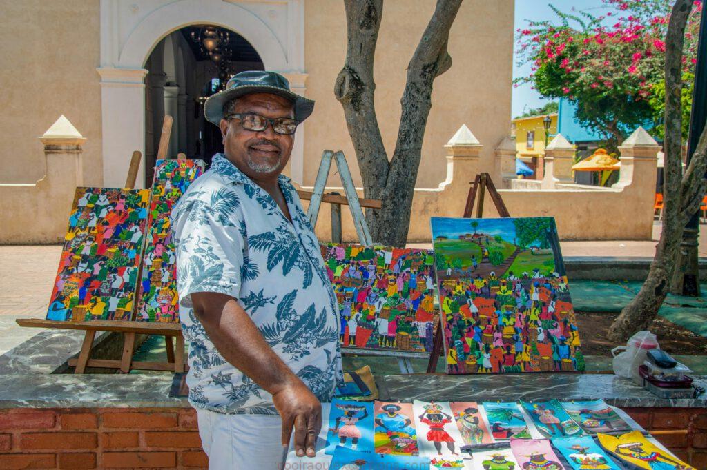 pexels photo 26551067 26551067 Local artist smiles at his colorful art stall in a sunny outdoor market setting.